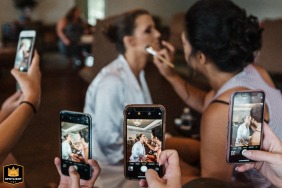 Bride in Magnolia Room, Rock Hill, SC gets makeup done, while being photographed by bridesmaids with their phones.